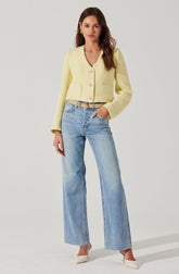 A woman is wearing a yellow, textured, long-sleeved, button-up cardigan with a v-neckline and two front pockets. She pairs it with high-waisted, light blue wide-leg jeans that have a slightly faded wash. She accessorizes with gold jewelry, including earrings and a layered necklace, and wears beige pointed-toe heels.